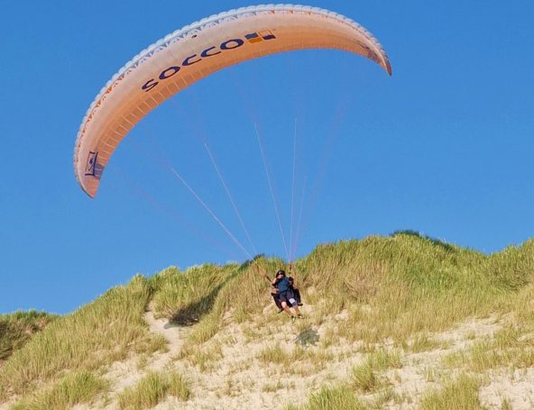 Paraglider schwebt über eine grüne Düne vor klarem, blauem Himmel.